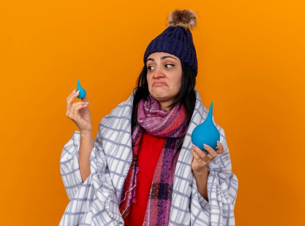 Confused young ill woman wearing winter hat and scarf wrapped in plaid holding enemas looking at little one isolated on orange wall