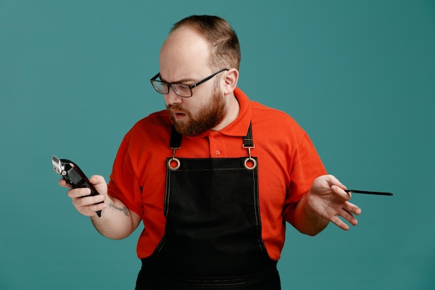 Free photo confused young male barber wearing glasses red shirt and barber apron holding hair clipper and teaser comb looking at clipper isolated on blue background