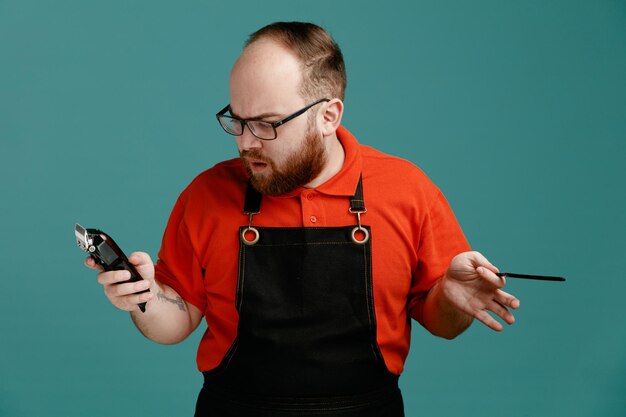 Confused young male barber wearing glasses red shirt and barber apron holding hair clipper and teaser comb looking at clipper isolated on blue background