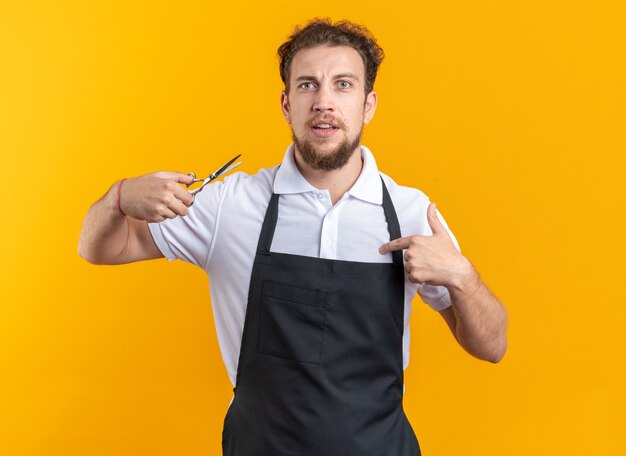 Confused young male barber wearing uniform holding scissors points at himself isolated on yellow wall