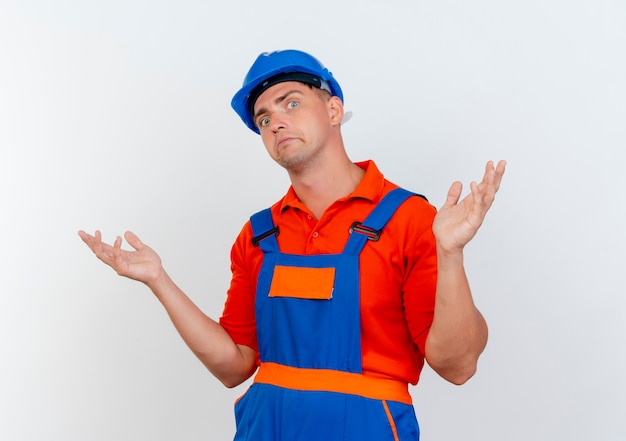 Confused young male builder wearing uniform and safety helmet spreads hands 