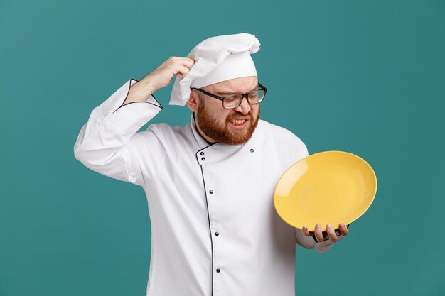 Confused young male chef wearing glasses uniform and cap holding and looking at empty plate keeping hand on head isolated on blue background