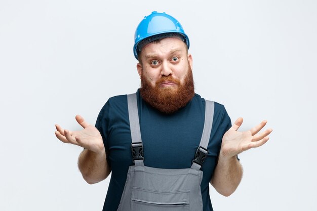 Confused young male construction worker wearing safety helmet and uniform looking at camera showing empty hands isolated on white background