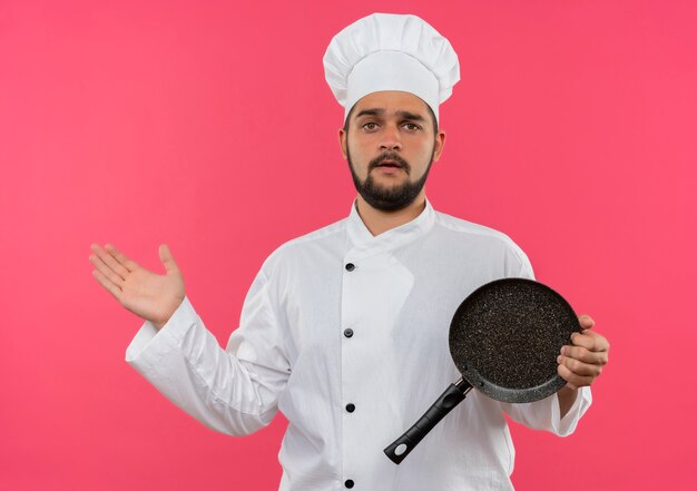 Confused young male cook in chef uniform holding frying pan and showing empty hand isolated on pink wall