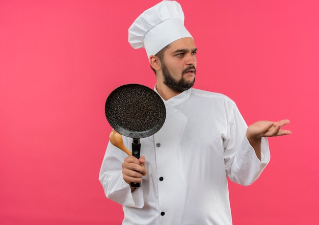 Смущенный молодой мужчина-повар в униформе шеф-повара держит сковороду и ложку, глядя в сторону и показывая пустую руку, изолированную на розовой стене с копией пространства