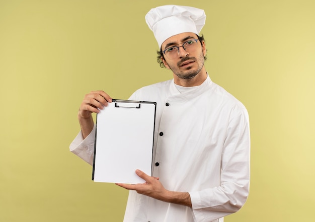 confused young male cook wearing chef uniform and glasses holding clipboard 