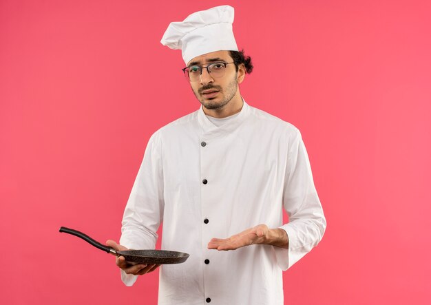 Confused young male cook wearing chef uniform and glasses holding and points with hand to frying pan 