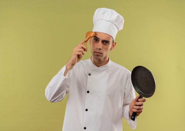  confused young male cook wearing chef uniform holding frying pan putting spoon on his forehead on isolated green wall