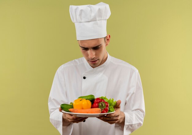 Confused young male cook wearing chef uniform looking at vegetables in his hand on isolated green wall with copy space