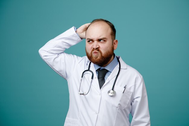 Confused young male doctor wearing medical coat and stethoscope around his neck looking at side scratching his head isolated on blue background