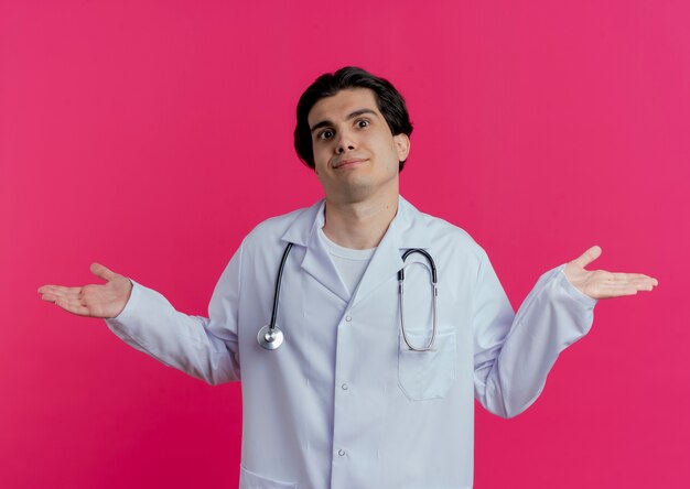 ピンクの壁に隔離された空の手を示す医療ローブと聴診器を身に着けている混乱した若い男性医師