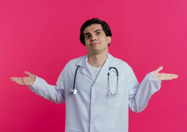 Confused young male doctor wearing medical robe and stethoscope  showing empty hands isolated on pink wall