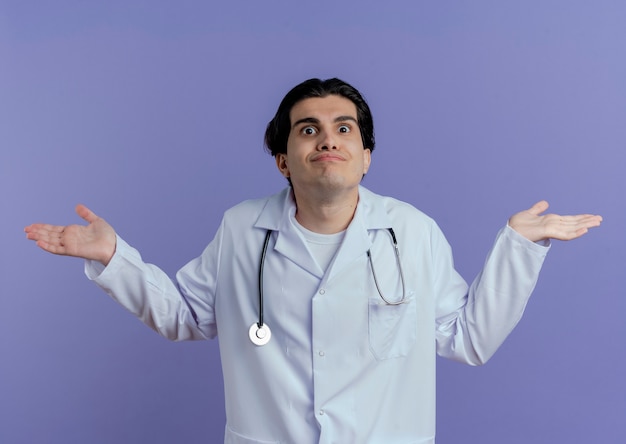 Confused young male doctor wearing medical robe and stethoscope  showing empty hands isolated on purple wall