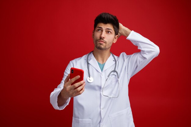 Giovane medico maschio confuso che indossa uniforme medica e stetoscopio intorno al collo che tiene il telefono cellulare tenendo la mano sulla parte posteriore della testa guardando in alto isolato su sfondo rosso