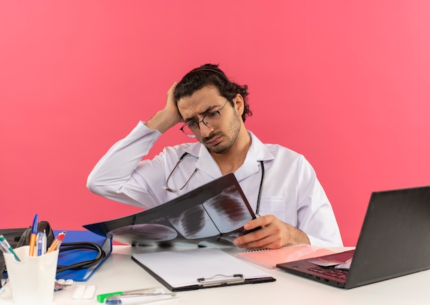 Confused young male doctor with medical glasses wearing medical robe with stethoscope sitting at desk