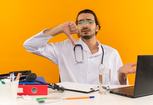 Смущенный молодой мужчина-врач в медицинских очках в медицинском халате со стетоскопом, сидя за столом