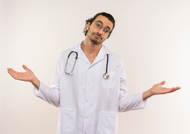 Confused young male doctor with optical glasses wearing white robe with stethoscope spreads hands