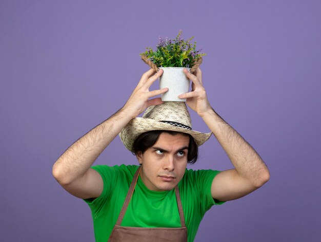 Confused young male gardener in uniform wearing gardening hat holding flower in flowerpot on head