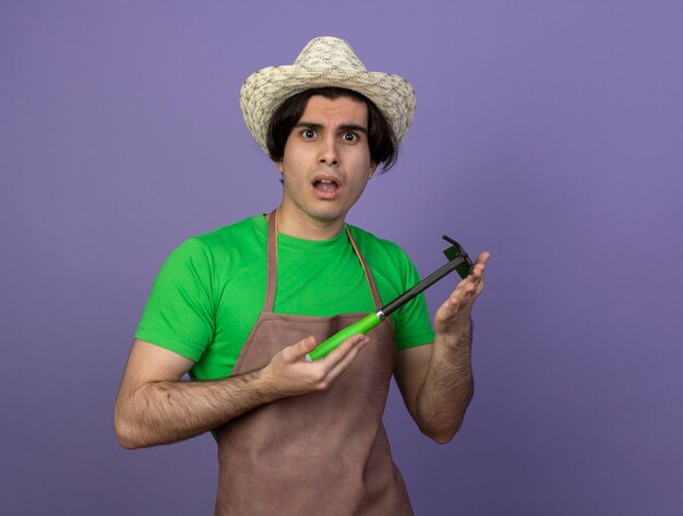 Confused young male gardener in uniform wearing gardening hat holding hoe rake