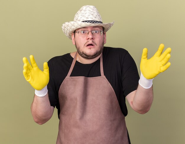 オリーブ グリーンの壁に手を広げてガーデニング帽子と手袋を身に着けている混乱した若い男性庭師