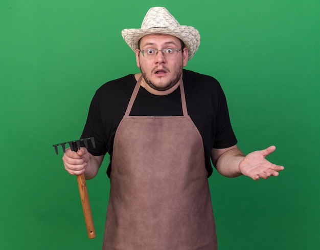 Confused young male gardener wearing gardening hat holding rake spreading hands isolated on green wall