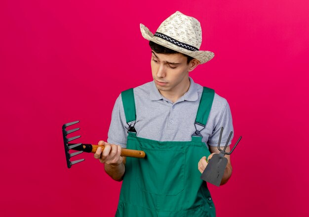 Confused young male gardener wearing gardening hat holds hoe rake and looks at rake