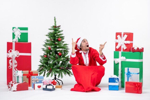 Confused young man dressed as Santa claus with gifts and decorated Christmas tree sitting on the ground looking above on white background