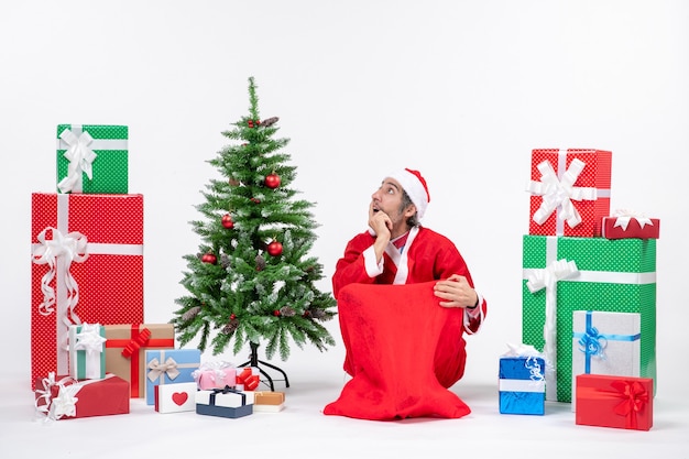 Confused young man dressed as Santa claus with gifts and decorated Christmas tree sitting on the ground on white background