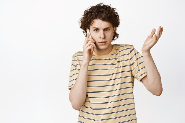 Confused young man having a strange phone call shrugging and looking puzzled talking on mobile standing over white background