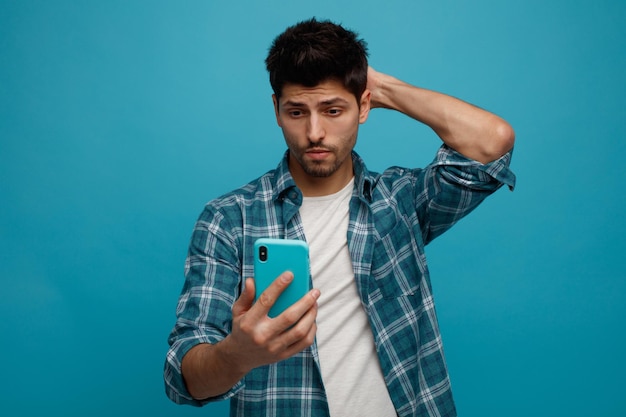 Смущенный молодой человек держит и смотрит на мобильный телефон, держа руку на затылке, изолированный на синем фоне Premium Фотографии