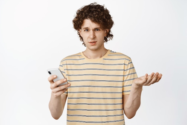 Confused young man holding phone reacting to smth strange staring frustrated and annoyed standing over white background