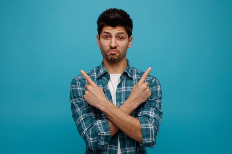 मुफ़्त फोटो confused young man keeping hands crossed looking at camera pointing to sides isolated on blue background with copy space