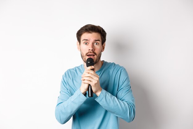 Смущенный молодой человек нервно смотрит в камеру во время пения караоке, держа микрофон, стоя на белом фоне.