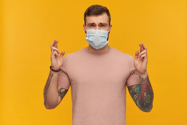 Confused young man in pink tshirt and virus protective mask on face against coronavirus with beard and tattoo keeps fingers crossed and making a wish over yellow wall