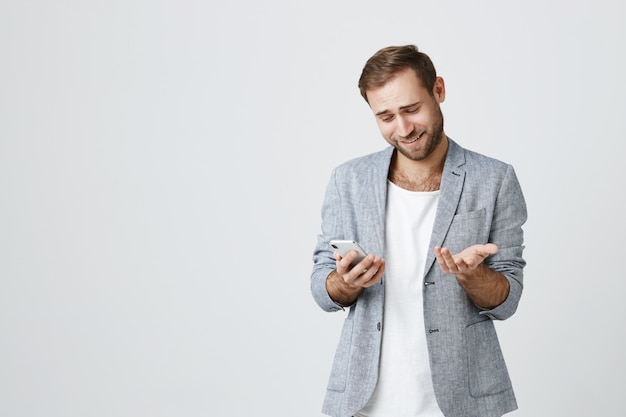 Confused young man shrugging at mobile phone screen, smiling