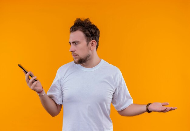 Смущенный молодой человек в белой футболке, глядя на телефон на руке на изолированной оранжевой стене