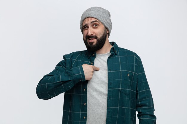 Confused young man wearing winter hat looking at camera pointing at himself isolated on white background