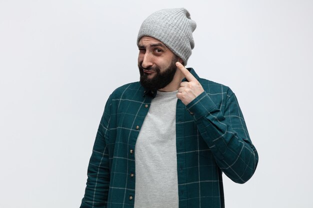 Confused young man wearing winter hat looking at camera pointing at himself isolated on white background