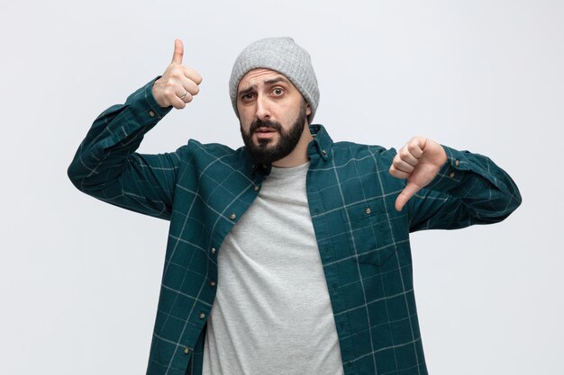 Confused young man wearing winter hat looking at camera showing thumbs up and down isolated on white background