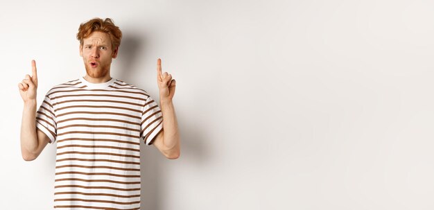 Confused young man with red curly hair pointing fingers up staring shocked and puzzled standing over