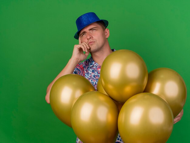 Confused young party guy wearing blue hat standing behind balloons and putting hand on cheek isolated on green