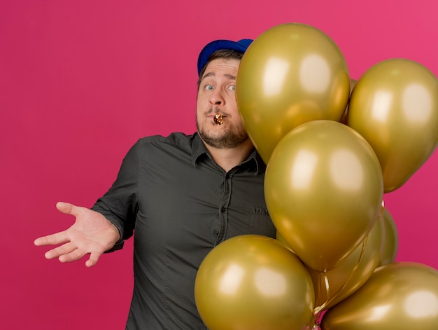 Confused young party guy wearing blue hat standing near balloons holding party blower in mouth spreading hands isolated on pink