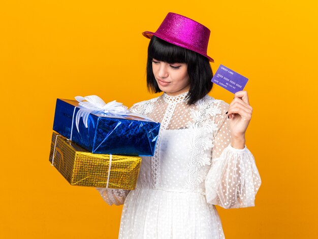 Confused young party woman wearing party hat holding gift packages and credit card looking at packages isolated on orange wall