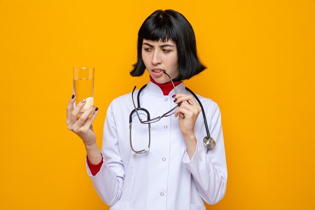 Confuso giovane bella ragazza caucasica in uniforme da medico con stetoscopio che tiene in mano un bicchiere d'acqua e occhiali ottici guardando a lato