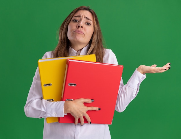 Confused young pretty caucasian girl holds file folders and keeps hand open on green 