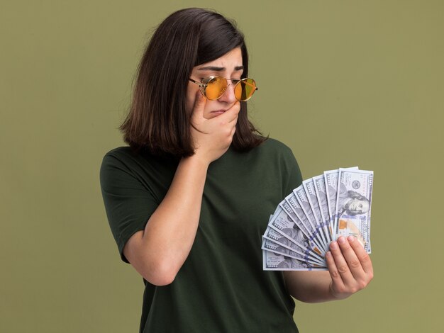 Confused young pretty caucasian girl in sun glasses puts hand on chin holding and looking at money on olive green