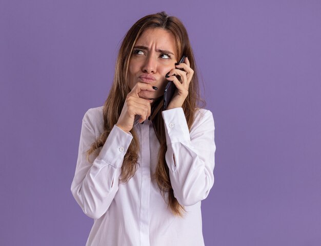 Смущенная молодая симпатичная кавказская девушка разговаривает по телефону и кладет руку на подбородок, глядя вверх изолированно на фиолетовой стене с копией пространства