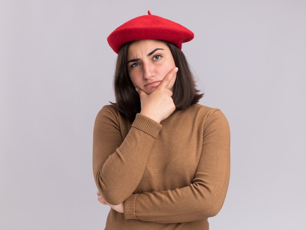 Confused young pretty caucasian girl with beret hat holds chin  isolated on white wall with copy space