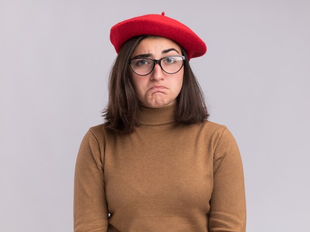 Confused young pretty caucasian girl with beret hat and in optical glasses looking at camera on white
