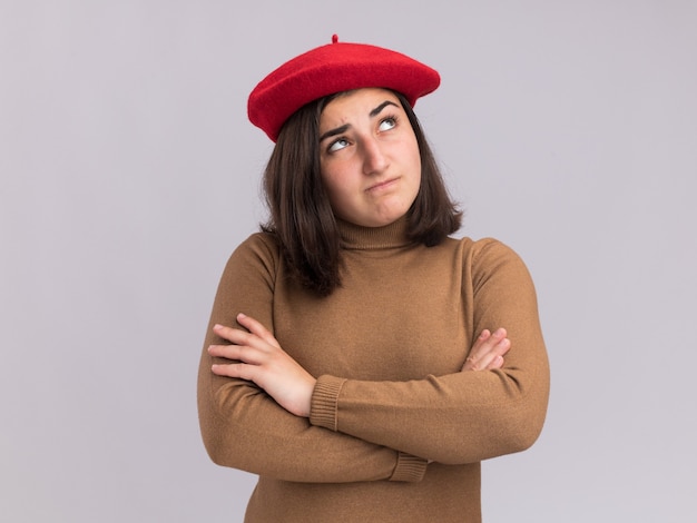 Confusa giovane ragazza abbastanza caucasica con cappello berretto sta con le braccia incrociate, alzando lo sguardo su bianco